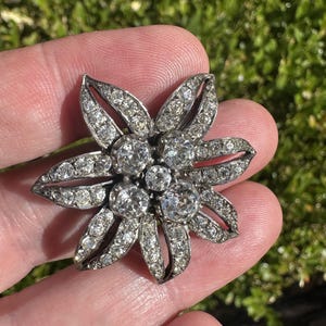 Pode incluir: Um broche em forma de flor de tom prateado com seis pétalas incrustadas com pequenas pedras transparentes e um centro de quatro pedras transparentes redondas e facetadas. A estrutura metálica tem um acabamento escuro e oxidado.