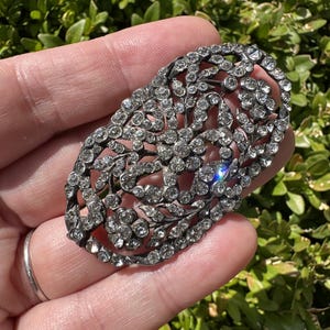 Op de afbeelding: Een ovale broche met een ingewikkeld opengewerkt metalen frame, versierd met talloze kleine, heldere, gefacetteerde strass-steentjes in een bloem- en rankpatroon. De metalen zetting heeft een donkere, geoxideerde afwerking die contrasteert.