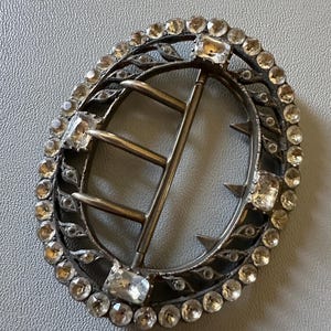 May include: An oval-shaped metal buckle featuring a dark, tarnished finish. The outer frame is decorated with a continuous border of small, clear, round faceted stones and four larger rectangular stones. A central metal bar includes three prongs.