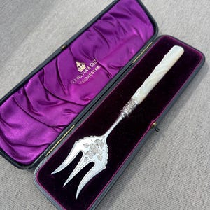 May include: An antique silver serving fork with a mother-of-pearl handle, displayed in a velvet-lined case. The fork features a floral design on the tines and a decorative silver collar. The case is black with a purple interior.
