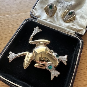 May include: A gold-toned frog brooch featuring clear rhinestones on its limbs and green cabochon eyes, accompanied by a pair of matching clip-on earrings with clear rhinestones and central green stones, all presented in a dark jewellery box.