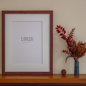 May include: A framed print with the text "LORIS FRAMING" in a decorative font. The frame is a dark reddish-brown colour. To the right is a small blue vase with dried flowers and a small, round, reddish-brown object.