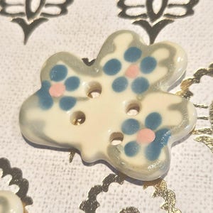 May include: A ceramic button in a clover shape, featuring a cream base with blue and pink floral patterns. The button has three holes for sewing and a gray border. The background has gold foil floral designs.
