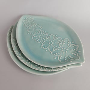 May include: A stack of three oval-shaped, light blue ceramic plates. Each plate features a white floral design along one side. The plates are stacked slightly offset, revealing the design on each.