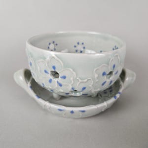 May include: A light gray ceramic bowl with a matching saucer, featuring raised white floral relief patterns and small blue painted dots. The bowl has cut-out centers in the flowers and sits on small feet, while the saucer has two side handles.