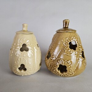 May include: Two ceramic decorative jars with lids. The jar on the left is a light cream color, while the jar on the right is a tan color. Both jars have a bulbous shape with floral cutouts and a small handle on the lid.