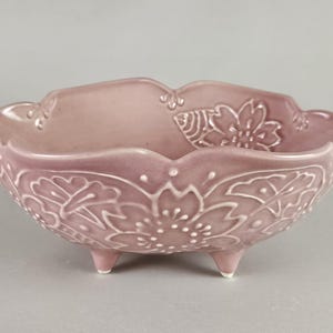 May include: A light purple ceramic bowl with a floral design. The bowl has a scalloped edge and sits on three small feet. The floral pattern is raised and detailed, adding texture to the piece. The bowl is likely used for decorative purposes.