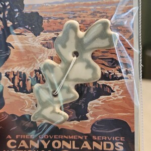 May include: A light green and white ceramic button with a unique, abstract shape. The button has two holes and is attached to a white string. The background features a vintage poster with the text "Canyonlands National Park".