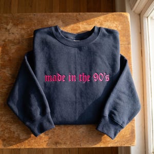 May include: A navy blue sweatshirt with the phrase "made in the 90's" embroidered in pink script across the chest. The sweatshirt is folded neatly on a wooden surface, with the sleeves tucked in.