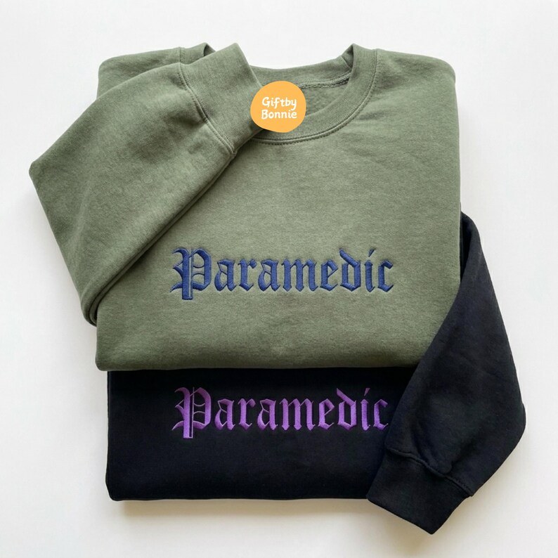 May include: An olive green sweatshirt with dark blue embroidered text reading "Paramedic" in a gothic font, layered over a black sweatshirt with purple embroidered text reading "Paramedic" in the same gothic font, featuring a small orange logo.