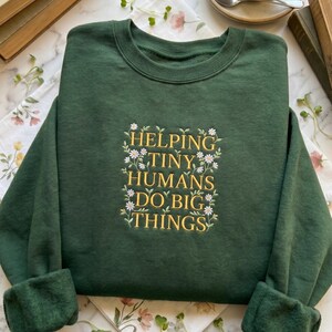 Peut inclure: Un sweat-shirt vert foncé avec l'inscription brodée en or "HELPING TINY HUMANS DO BIG THINGS", entourée de petites fleurs blanches. Le sweat-shirt a des manches longues et un col rond.