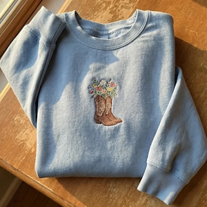 Puede incluir: Sudadera azul claro con cuello redondo y puños acanalados. Presenta un diseño bordado de una bota de vaquero marrón llena de flores coloridas. La sudadera está doblada sobre una superficie de madera.