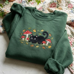 May include: Dark green sweatshirt with an embroidered design of a black cat amongst red-spotted mushrooms and autumn leaves. The crew neck sweatshirt has long sleeves and a relaxed fit, suitable for casual wear.