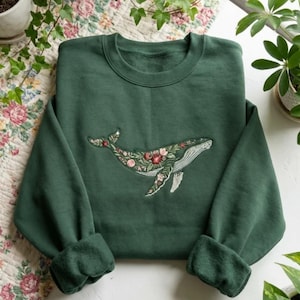 以下が含まれることがあります： ピンク、赤、緑の色合いの花で埋められた刺繍されたクジラのデザインが特徴の濃い緑色のスウェットシャツ。クジラは白いアクセントで詳細に描かれています。スウェットシャツはクルーネックで、袖口付きの長袖です。