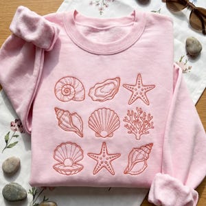 May include: A light pink sweatshirt featuring embroidered coral-colored seashells, starfish, and coral. The design includes a variety of sea life, such as a spiral shell, oyster, and a clam with a pearl. The sweatshirt is folded on a floral patterned cloth.