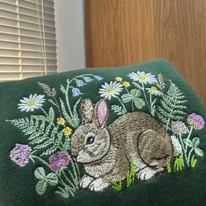 May include: A dark green embroidered cushion featuring a detailed design of a brown rabbit surrounded by wildflowers, including daisies, clover, and ferns. The embroidery is in shades of white, yellow, green, and purple.