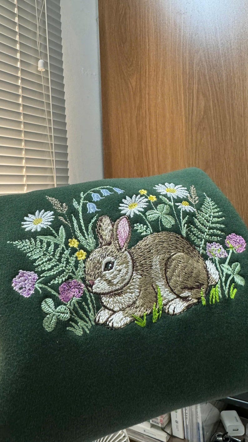 May include: A dark green embroidered cushion featuring a detailed design of a brown rabbit surrounded by wildflowers, including daisies, clover, and ferns. The embroidery is in shades of white, yellow, green, and purple.