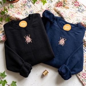 May include: A black sweatshirt and a navy blue sweatshirt, each featuring a small embroidered beetle with pink and green floral details on the chest. Both garments have a round neckline and long sleeves. Each includes a round orange Giftby Bonnie sticker.