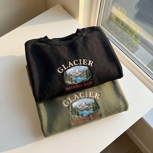 Op de afbeelding: Twee sweatshirts, één zwart en één olijfgroen, zijn op elkaar gestapeld. Elk heeft een geborduurd ontwerp met het woord "GLACIER" boven een landschapsbeeld van bergen, een meer en bomen, met "NATIONAL PARK" eronder.