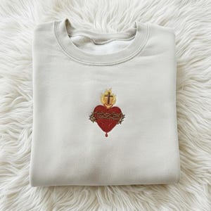 Puede incluir: Sudadera beige claro con cuello redondo. La parte delantera presenta un diseño bordado de un corazón rojo rodeado por una corona de espinas, con una cruz encima y una llama. El diseño está centrado en el pecho.