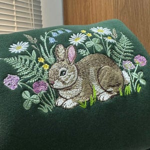 May include: A dark green embroidered fabric featuring a detailed depiction of a brown rabbit amidst wildflowers and foliage. The embroidery includes daisies, clover, and ferns, creating a nature-themed design.