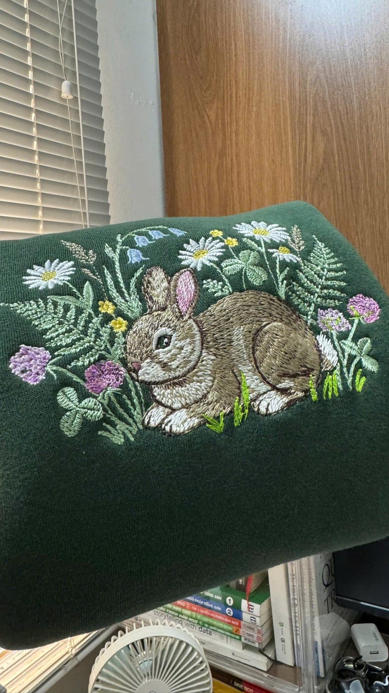 May include: A dark green embroidered fabric featuring a detailed depiction of a brown rabbit amidst wildflowers and foliage. The embroidery includes daisies, clover, and ferns, creating a nature-themed design.