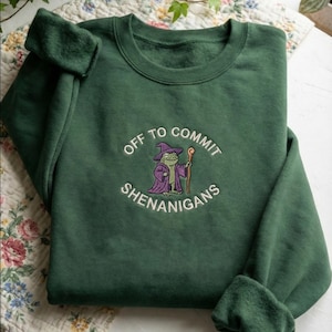 May include: A dark green sweatshirt with the embroidered phrase "OFF TO COMMIT SHENANIGANS" and a cartoon frog wearing a wizard hat and robe. The frog is holding a staff. The sweatshirt has a crew neck and long sleeves.