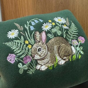 May include: Dark green sweatshirt with an embroidered design featuring a brown rabbit surrounded by wildflowers, including daisies, clover, and bluebells. The embroidery is detailed and colorful, creating a nature-inspired theme.