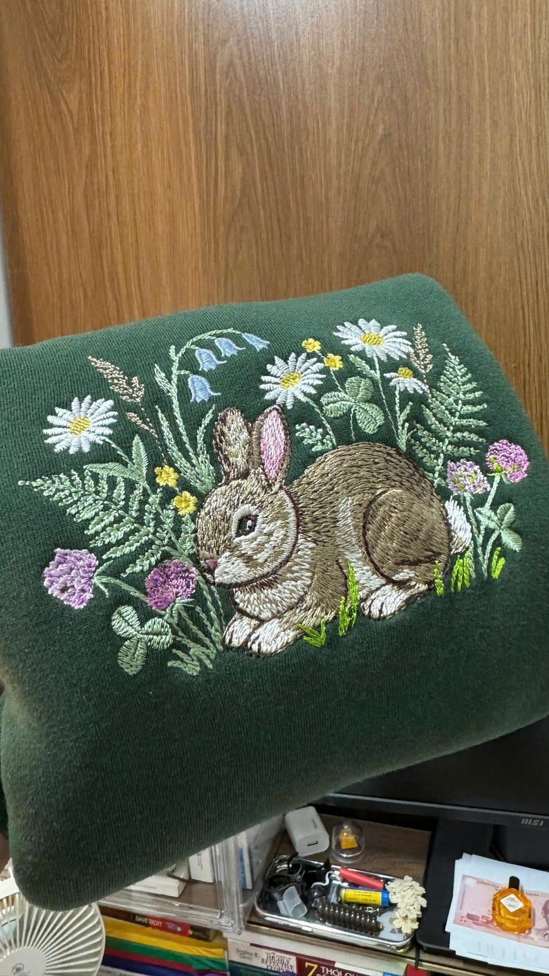 May include: Dark green sweatshirt with an embroidered design featuring a brown rabbit surrounded by wildflowers, including daisies, clover, and bluebells. The embroidery is detailed and colorful, creating a nature-inspired theme.