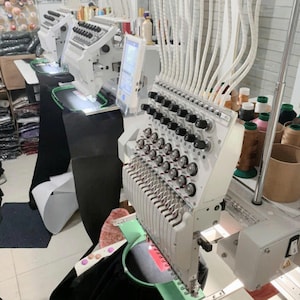 May include: A multi-needle industrial embroidery machine in white with numerous thread spools, tension dials, and a digital display screen. The machine features a green circular hoop holding black fabric, with white tubes feeding thread to the needles.