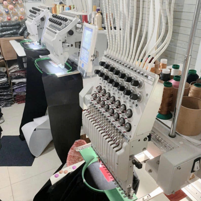 May include: A multi-needle industrial embroidery machine in white with numerous thread spools, tension dials, and a digital display screen. The machine features a green circular hoop holding black fabric, with white tubes feeding thread to the needles.