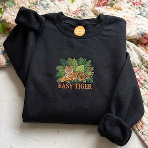 Puede incluir: Una sudadera negra de cuello redondo con un diseño bordado de un tigre naranja y negro descansando entre hojas tropicales verdes. Debajo del tigre, el texto EASY TIGER está bordado en hilo naranja. Un pequeño logotipo circular naranja dice Giftby Bonnie.