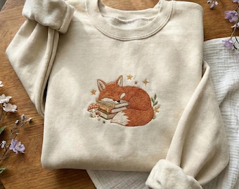 May include: Cream-colored sweatshirt featuring an embroidered design of a sleeping fox curled up on a stack of books. The fox is orange and white, with small stars and mushrooms around it. The sleeves are rolled up.