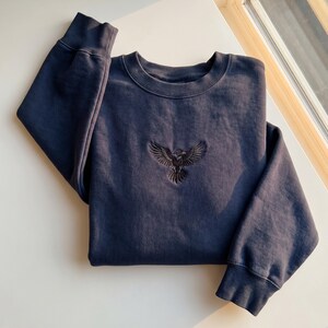 Könnte beinhalten: Ein marineblaues Sweatshirt mit Rundhalsausschnitt und langen Ärmeln. Das Sweatshirt hat ein gesticktes schwarzes Adlerdesign auf der Brust. Das Kleidungsstück ist sauber auf einer weißen Oberfläche gefaltet.