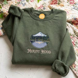 Może przedstawiać: Oliwkowa bluza z okrągłym dekoltem i haftem przedstawiającym ośnieżone pasmo górskie nad jeziorem otoczonym sosnami. Napis "MOUNT HOOD" jest wyhaftowany w jasnym kremowym kolorze pod wzorem górskiego krajobrazu.