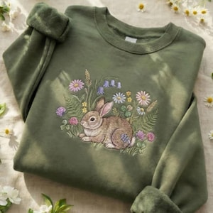 May include: Olive green sweatshirt featuring an embroidered design of a brown rabbit surrounded by colorful wildflowers. The design includes daisies, bluebells, and clover. The sweatshirt has a crew neck and long sleeves with rolled cuffs. A cozy and nature-inspired apparel item.