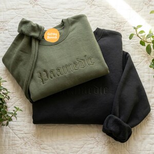 May include: An olive green crewneck sweatshirt with the word Paamedic embroidered in a gothic font on the chest, layered over a black crewneck sweatshirt with the same word embroidered in black thread. A small yellow circular logo reads Giftby Bonnie.