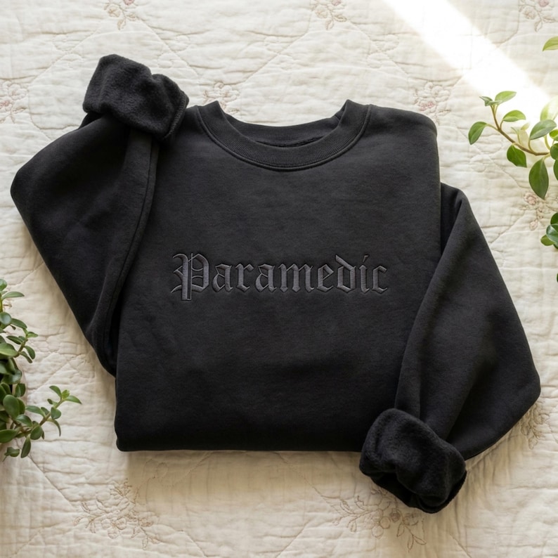 May include: A black long-sleeve sweatshirt featuring the word "Paramedic" embroidered in a matching black thread across the chest. The garment has a crew neckline, ribbed cuffs, and a soft, fleece-lined interior visible at the sleeve openings.