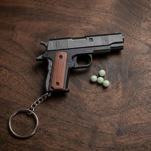 以下が含まれることがあります： 茶色のテクスチャ加工されたグリップパネルと銀色の金属リングを備えた、黒いミニチュアピストルのキーホルダー。ピストルの横には、4つの小さな淡い緑色の球状のペレットが配置されています。このオブジェクトは、リアルなスライド、トリガー、ハンマーのデザインが特徴です。