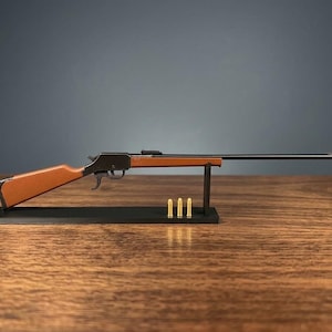 May include: A miniature lever-action rifle model featuring a black metal barrel and receiver with a brown stock and forearm. The rifle is mounted on a black rectangular display stand, accompanied by three small brass-colored cartridges.