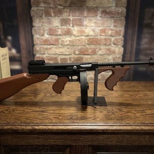May include: A black and brown replica submachine gun featuring a prominent circular drum magazine, a long barrel with an orange tip, and wooden-toned stock and grips. The firearm is mounted on a metal stand, showcasing its mechanical design.