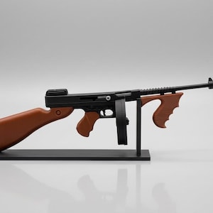 May include: A black and brown model of a submachine gun featuring a long barrel, a circular drum magazine, a wooden-colored stock, and a matching front grip. The model is mounted on a black rectangular base with a vertical support stand.