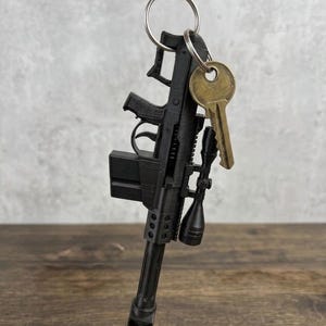 May include: A black 3D-printed rifle keychain featuring a detailed scope, a box magazine, and a muzzle brake. The rifle model is attached to a silver metal split ring that also holds a single brass-colored house key, creating a functional accessory.