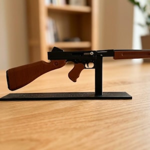 Può includere: Una replica in miniatura di un mitra Thompson con telaio in metallo nero, calcio, impugnatura a pistola e astina con texture in legno marrone. Il modello è montato su un supporto rettangolare nero, accompagnato da un caricatore nero separato.