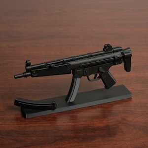 Könnte beinhalten: Ein schwarzes Miniaturmodell einer MP5-Maschinenpistole, montiert auf einem rechteckigen schwarzen Präsentationsständer. Das Modell verfügt über einen kurzen Lauf, einen Pistolengriff, einen Abzugsbügel und ein Visier. Ein separates gebogenes Magazin liegt auf der Basis.