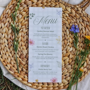 May include: A printed menu card with a checkered background, floral accents, and the word "Menu" at the top. The menu lists starter, main, and dessert options, including Garden Harvest Salad and Petite Berry Tart. The card is placed on a woven basket.