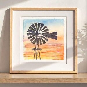 May include: Watercolor painting of a windmill set against a sunset sky. The windmill is black, and the sky transitions from blue to orange and yellow. The artwork is framed in a light wood frame.