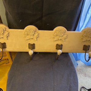 Może przedstawiać: Kremowy, drewniany wieszak ścienny z czterema haczykami. Wieszak posiada rzeźbione wzory róż i dekoracyjne metalowe i ceramiczne haczyki. Element w stylu vintage do wieszania płaszczy, czapek lub akcesoriów.