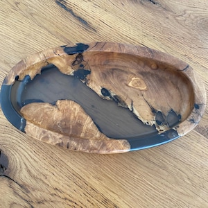 Può includere: Una ciotola ovale in legno caratterizzata da venature naturali color marrone chiaro con nodi e crepe visibili riempiti da un inserto in resina grigio scuro liscia che crea un motivo a contrasto simile a un fiume al centro del piatto.