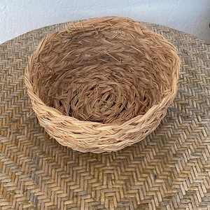 Peut inclure: Un panier rond et peu profond tressé à partir d'herbes ou de roseaux séchés de couleur brun clair naturel. Le panier présente une construction enroulée et texturée avec des brins visibles formant un bol robuste à sommet ouvert.